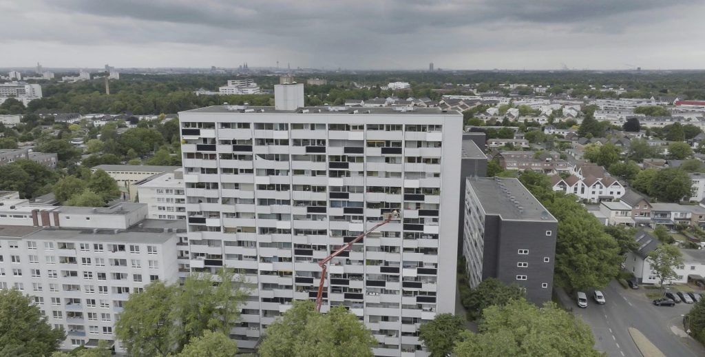 Luftaufnahme eines Hochhauses in der Robert-Schumann-Straße im Kölner Stadtteil Neubrück, Teil des KlimaVeedel-Projekts der RheinEnergie. Ein großer Montagekran installiert zahlreiche Balkonkraftwerke (Mini-Solaranlagen) an der Fassade des Gebäudes. Die Solarmodule werden auf den Balkonen angebracht, um klimafreundliche Energie direkt vor Ort zu erzeugen. Im Hintergrund erstreckt sich die dichte Bebauung von Köln mit viel Grün und einer Mischung aus Einfamilienhäusern und Wohnanlagen. Dieses Vorzeigeprojekt fördert die Energiewende im Quartier, steigert die Energieautarkie der Mieter*innen und macht Photovoltaik im urbanen Raum sichtbar und nutzbar. Wichtige Begriffe für SEO: Balkonkraftwerke Köln, RheinEnergie KlimaVeedel, Solaranlage Balkon Neubrück, nachhaltige Stadtentwicklung Köln, Photovoltaik Wohngebäude, Energiewende im Veedel.